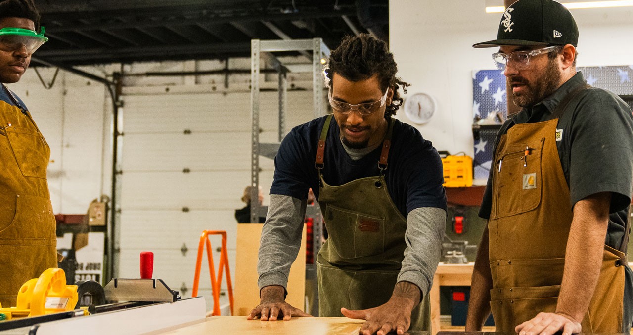 Teacher oversees student cutting wood with table saw in workshop
