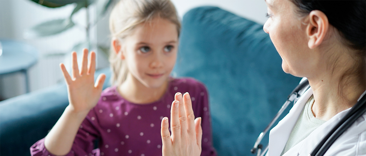 A woman pediatrician interacts with a young female patient who looks tentative but smiles. 